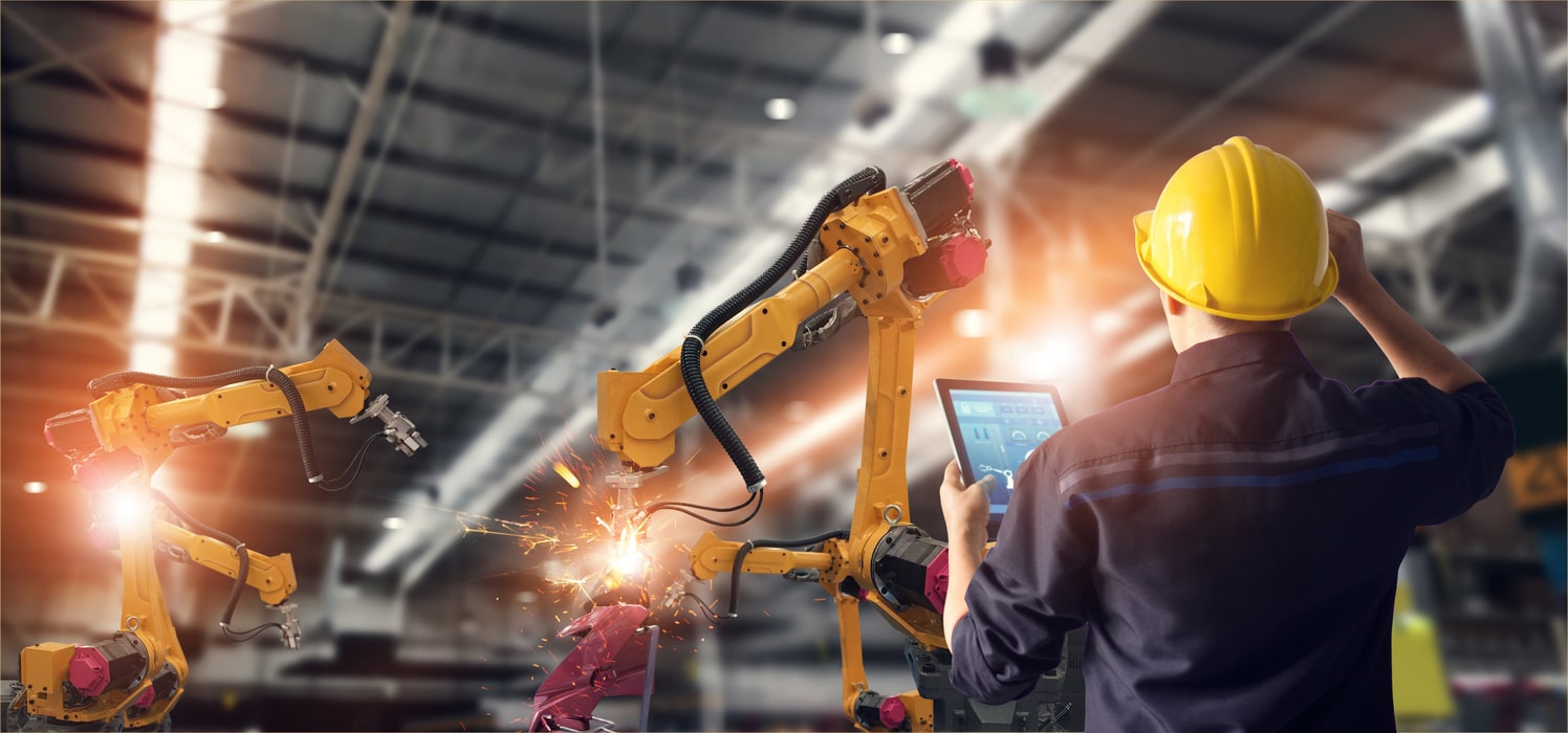 A Worker In A Hard Hat Supervises Two Robotic Arms That Are Welding Parts Together.