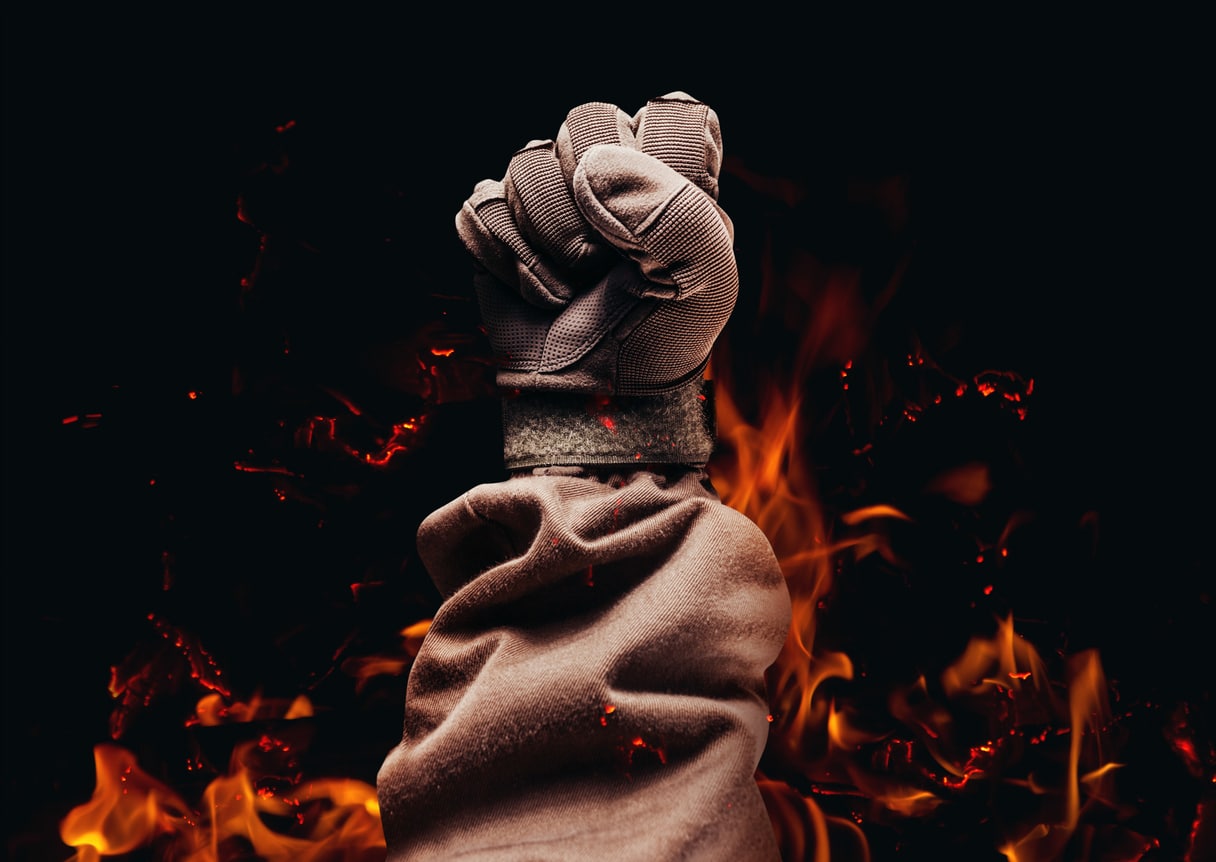 A Raised Fist Wearing A Fire-resistant Glove And Jacket With Flames In The Background.